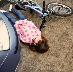 Little girl knocked off bike, lying dead by car, eyes open. She is not wearing a helmet.