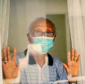 Senior man looking outside the window of a nursing home while wearing mask