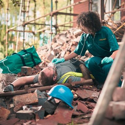 a builder or roofer has fallen from the top elevation of scaffold on a construction site and landed on rubble at the base of the building . He is grimacing in pain . A paramedic tends to him.