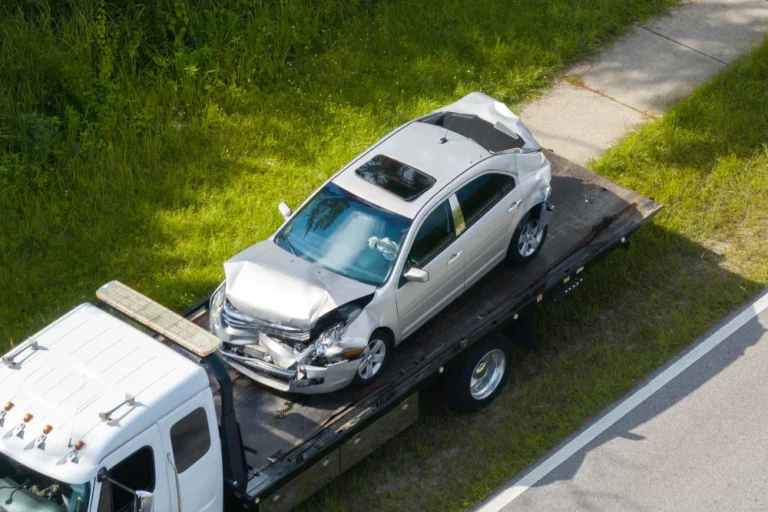 Grey car with front end and rear end damage on a flat bed tow truck