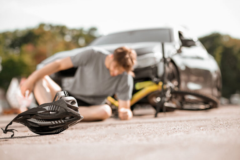 Man sitting on the pavement leaning to his side with a damaged bike and parked car in the background.