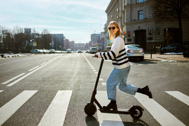 Woman riding e-scooter across a city street crosswalk.